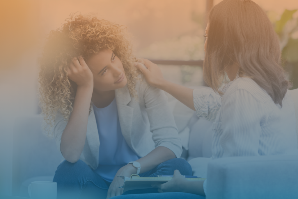 One woman places her hand on another woman's shoulder while sitting facing each other