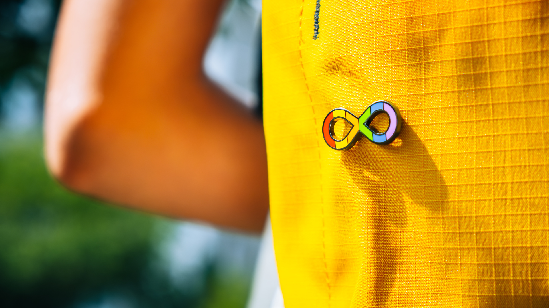 A close up photo of a person wearing an infinity rainbow pin on their yellow backpack